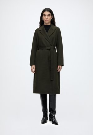 Femme aux longs cheveux foncés portant un manteau vert foncé ceinturé et des bottes en cuir noires montantes jusqu'aux genoux sur un fond blanc uni.