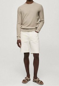 Mann trägt einen beigen, gerippten Pullover mit langen Ärmeln, helle Shorts und beigen Sandalen mit Kreuzgurt, steht vor einem schlichten hellen Hintergrund.