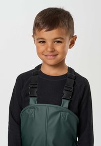Jeune garçon souriant aux cheveux bruns courts porte un haut noir à manches longues et une salopette verte avec des bretelles noires ajustables, tourné vers l’avant.