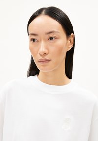 Vit bomullströja med rund hals och en texturerad, ljus broderidetalj på framsidan. Enkel och minimalistisk design.