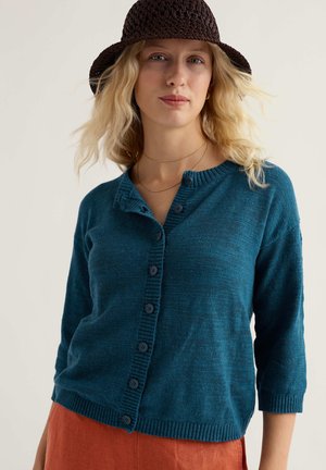 Sieviete ar viļņainiem blondiem matiem, tērpusies tumši brūnā adītā cepurē, tirkīzzilā pogātas kardiganā un rūsas krāsas biksēs uz vienkārša gaiša fona.