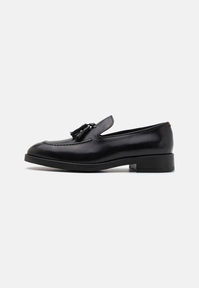 Mocassins en cuir noir avec des accents à franges, détails cousus, bout rond et un talon bas empilé. Fini lisse avec un design épuré.