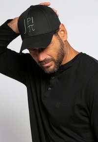 Schwarze Kappe mit gesticktem Logo, Gewebestruktur sichtbar. Tragen Sie sie mit einem schwarzen Langarmshirt mit Knöpfen und dezentem Logo.