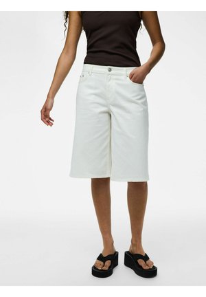 Femme portant un short blanc mi-genou, un top noir sans manches et des sandales plateformes noires, debout avec une main dans la poche.