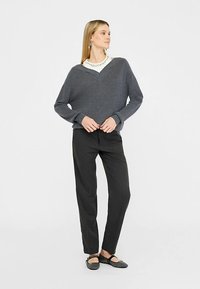 Sivi pleteni džemper s V-izrezom, uparen s crnim hlačama na pruge. Model nosi crne ravne cipele, prikazujući opuštenu siluetu i teksturu.