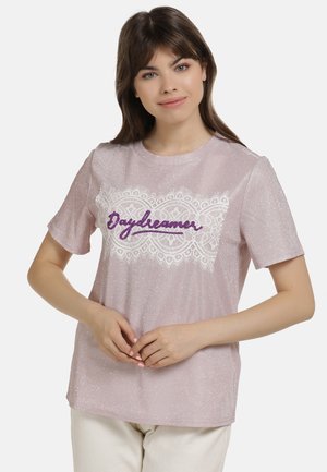Junge Frau trägt ein glänzendes hellrosa T-Shirt mit weißen Spitzendetails und dem vorn in Lila bestickten Wort „Daydreamer“.