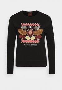 Sudadera negra con un colorido diseño floral bordado que lleva la palabra "WANDERER" y "Maison Scotch" en la parte inferior. Mangas largas, cuello redondo.