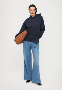 Woman wearing a navy hoodie and wide-leg blue jeans, holding a large brown leather bag, standing against a plain white background.