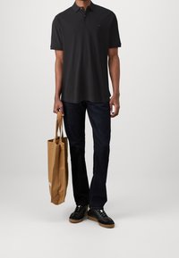 Polo noir à manches courtes, jeans bleu foncé et baskets noires. Tient un sac fourre-tout beige. Design minimaliste, sans motifs visibles.