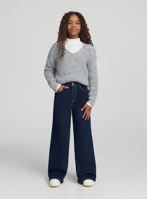 Maglione grigio lavorato a maglia con scollo a V, abbinato a jeans blu scuro a gamba larga. Dolcevita bianco casual sotto e sneakers bianche.