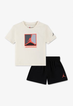 Krém színű, rövid ujjú Jordan póló narancssárga logóval és fekete rövidnadrág, amelynek bal lábán kis narancssárga Jordan logó található.