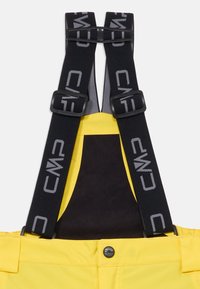 Keltaiset vedenpitävät haalarit, joissa on mustat säädettävät hihnat ja harmaat yksityiskohdat. Vyössä metallinen nappisuljin. Liukas pinta.