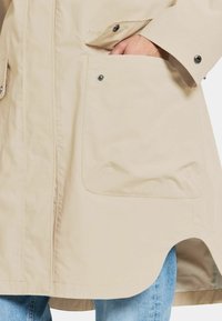 Beige regenjas met een rechte snit, grote voorkruiken, drukknoopsluitingen en een gebogen zoom. Gladde textuur en lichtgewicht stof.