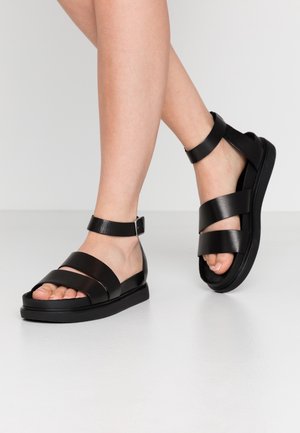 Sorte lædersandaletter har tre brede stropper hen over toppen, en ankelrem med spænde og en flad gummisål.