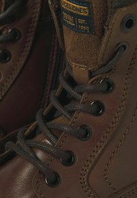 Las botas de cuero marrón presentan ojales de metal, cordones oscuros y una lengüeta de tela texturizada, con una pequeña etiqueta de logo y detalles de costura visibles.