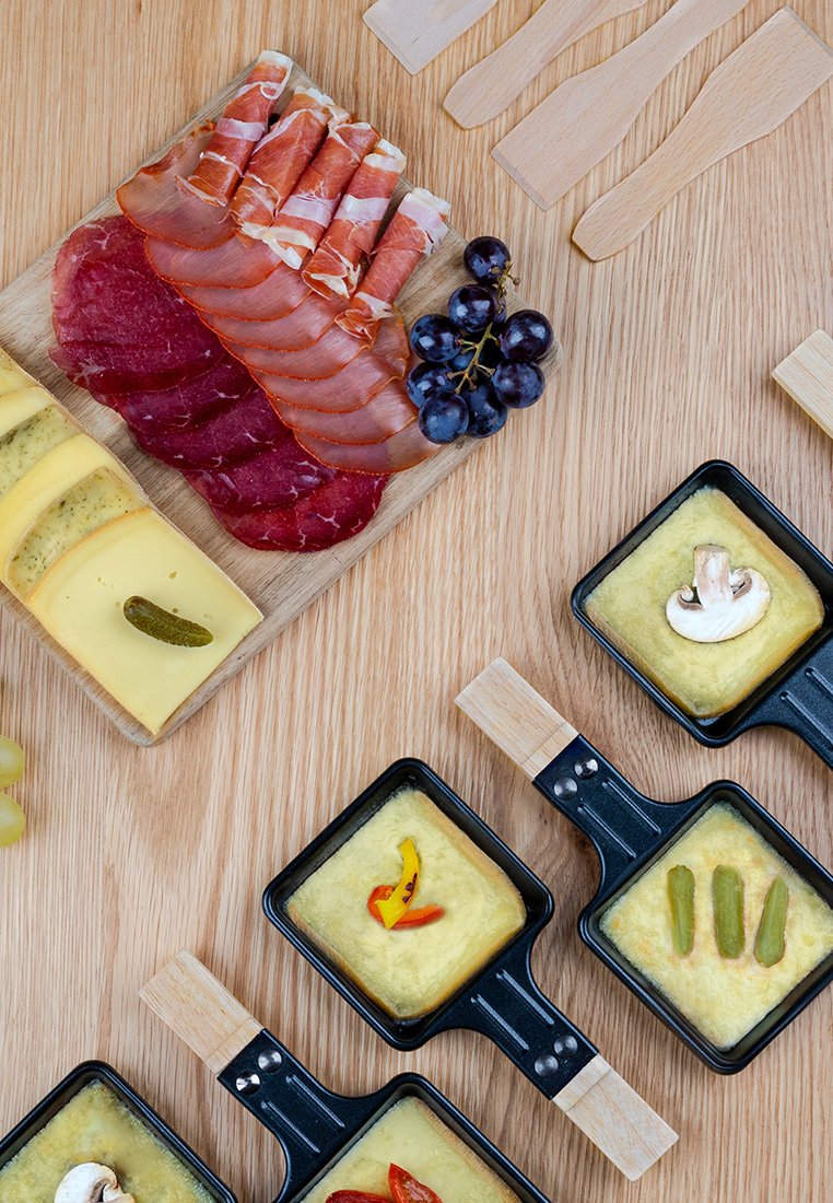 Viandes séchées tranchées, fromage et raisins sur une planche en bois avec de petites casseroles de fromage fondu surmontées de champignons, poivrons et cornichons.