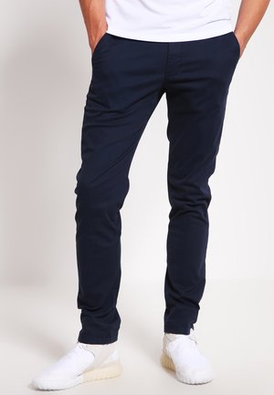 Chino navy blue dintr-un material neted, cu croială slim, buzunare laterale și un design frontal plat, asortat cu adidași albi.