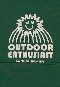 Sol sorridente com raios acima do texto "ENTUSIASTA DE ATIVIDADES AO AR LIVRE" e logotipo da Quiksilver em tecido verde escuro.