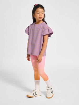 Junges Mädchen mit langen dunklen Haaren, das ein lila Top mit flatternden Ärmeln, pink-orange Farbverlauf-Leggings, weiße Socken und weiße Turnschuhe trägt und steht.