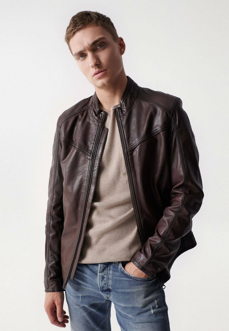 Braune Lederjacke mit Stehkragen, Reißverschluss und tailliertem Schnitt. Getragen über einem beigen, strukturierten Hemd und kombiniert mit blauen Jeans.