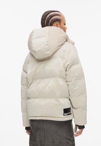 Pufferjack in lichtbeige met een gewatteerd ontwerp, glanzende textuur, een capuchon en een zwart labelaccent aan de zoom.
