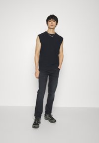 Maglietta nera senza maniche, jeans skinny scuri e scarpe da ginnastica nere. Design semplice, tessuto texturizzato, con una silhouette aderente.