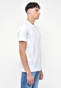 T-shirt bianca a girocollo in tessuto di cotone, maniche corte, vestibilità comoda. Indossata con jeans in denim blu, mostra un profilo laterale.