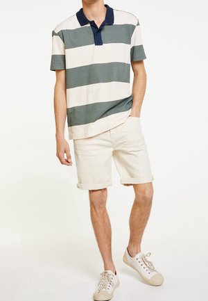 Mand iført grøn- og hvidstribet kortærmet polo-shirt, beige opkørte shorts og hvide sneakers, stående med den ene hånd i lommen.