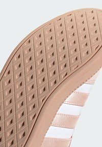 Semelle de chaussure en caoutchouc rose pâle avec un motif de grip texturé, arborant des rayures blanches sur le côté et une finition lisse sur le bord latéral.