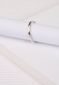 Zilveren bandring met een glad, glanzend oppervlak. Ronde, minimalistische vorm rustend op een wit cilindrisch voorwerp en een gestructureerde achtergrond.