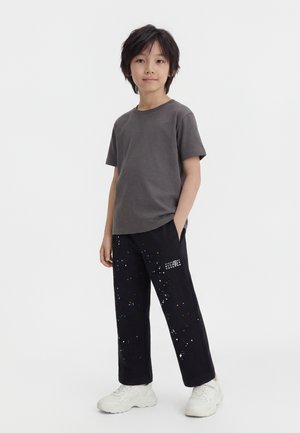 Barn med kort sort hår står med hænderne i lommerne iført en grå t-shirt, sorte bukser med malingssprøjt og hvide sneakers.