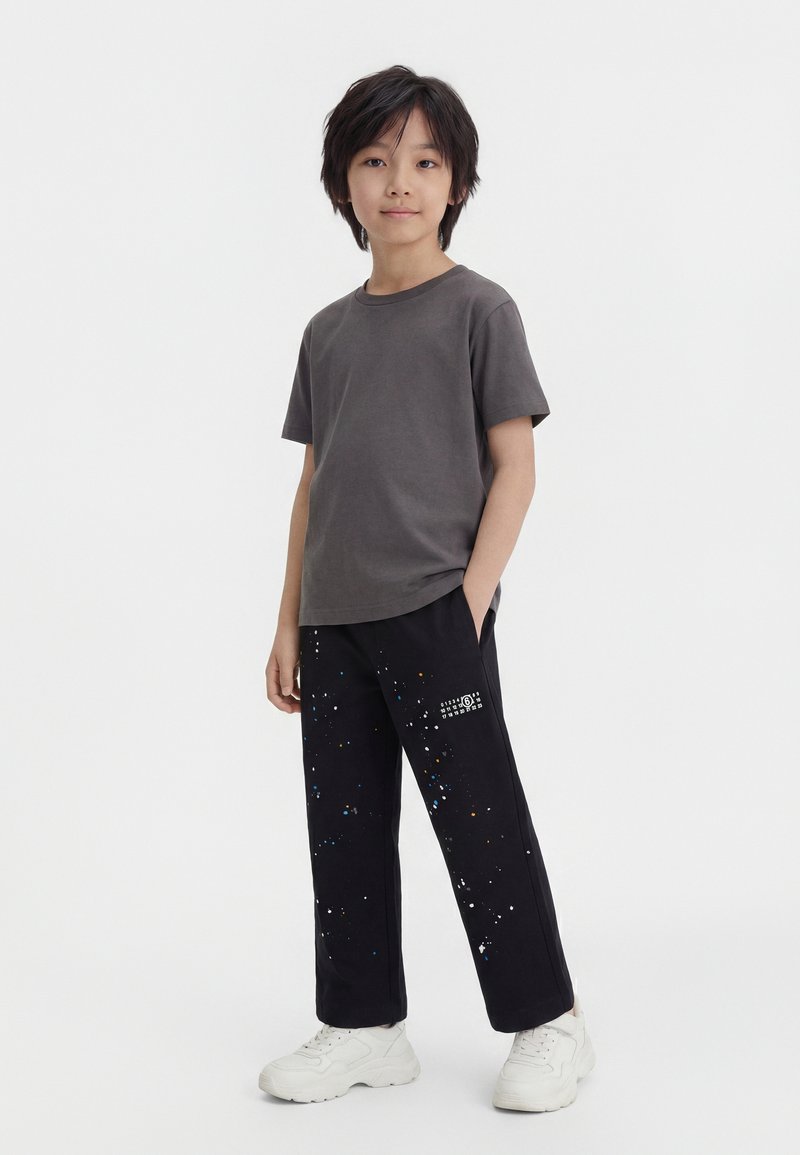 Παιδί με κοντά μαύρα μαλλιά στέκεται με τα χέρια στις τσέπες, φορώντας γκρι μπλουζάκι, μαύρο παντελόνι με πιτσιλιές μπογιάς και λευκά αθλητικά παπούτσια.