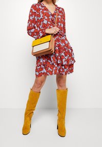 Vestido rojo con estampado floral y un dobladillo con volantes, combinado con un bolso de hombro amarillo y marrón y botas de ante amarillas hasta la rodilla.