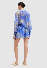 Majica s plavim i žutim uzorkom, lagana, teksturirana, s opuštenim krojem i balon rukavima, uparena s odgovarajućim kratkim hlačama, nosi se s laganim sandalama.