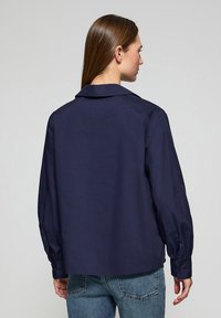 Camicia blu navy con colletto, maniche lunghe e polsini abbotttonati. Realizzata in tessuto liscio; presenta un taglio classico e una minimale decorazione.