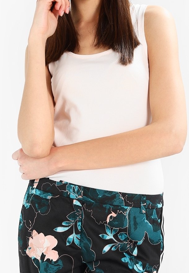 Femme portant un haut blanc sans manches et un pantalon noir avec des motifs floraux bleus et roses, posant avec un bras croisé et l'autre main près du visage.