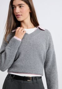 Szary, krótki sweter z miękkiego dzianiny z dekoltem w serek i prążkowanymi mankietami; nałożony na biały T-shirt z różową koszulą pod spodem.