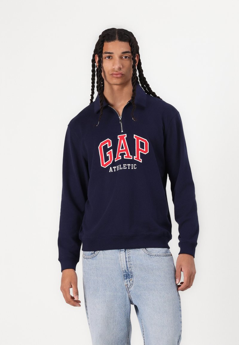 Sudadera azul marino con cremallera, con el texto rojo "GAP" y "ATHLETIC" debajo. Fabricada con un tejido suave, con puños y dobladillo acanalados.