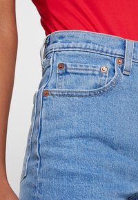 Jeans de tiro alto en azul claro con herrajes de cobre, diseño de cinco bolsillos y una textura de denim suave. Costuras visibles a lo largo de los bordes.