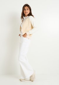 Cárdigan beige claro y blanco con una textura suave, que presenta un cierre de botones y puños de canalé en contraste, combinado con jeans acampanados blancos y zapatillas.