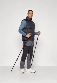 Černá prošívaná vesta přes modré tričko s dlouhým rukávem, doplněná šedými a černými kalhotami. Jedinec má v ruce trekingové hole a nosí bílé tenisky.