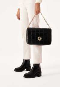 Persona che indossa pantaloni bianchi e stivali neri alla caviglia, mentre tiene una borsa a tracolla nera imbottita con catena dorata e logo circolare dorato.