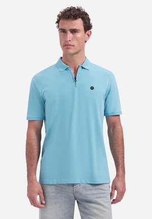 Man draagt een lichtblauw poloshirt met korte mouwen, ritssluiting en een zeshoekig logo op de borst, gecombineerd met lichtgewassen jeans, staand tegen een witte achtergrond.