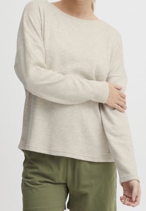 Kobieta ubrana w beżowy sweter z długim rękawem i oliwkowo zielone szorty, trzymająca lewą rękę prawą dłonią.