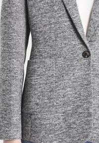 Blazer gris tissé avec un col cranté, une fermeture à un bouton et une poche avant. Tissu texturé et design structuré.