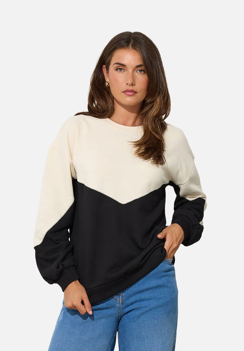 Woman with brown hair wearing a black and cream long-sleeve sweater and blue jeans, standing with one hand in her pocket.