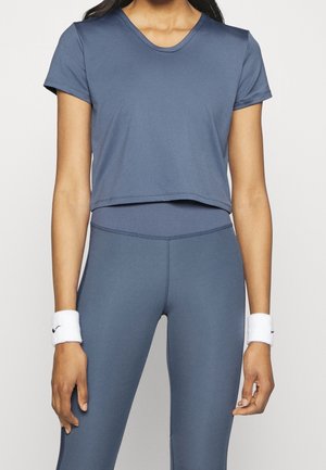 Kvinde iført en blå, kortærmet crop-top og matchende høj taljeleggings, med hvide håndledsbind, der har et lille sort logo.