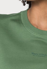 Primo piano di una persona che indossa una maglietta verde a girocollo con del testo blu scuro parzialmente visibile vicino al petto.