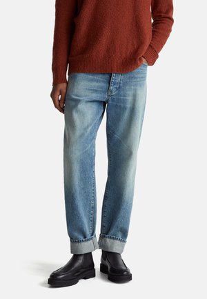 Vaqueros de mezclilla azul claro con un corte relajado, que presentan un dobladillo vuelto. Combinados con botas de piel negras hasta el tobillo y un suéter de textura rojo oscuro.