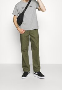 Homme portant un t-shirt Levi's gris, un pantalon vert olive, des baskets noires Nike et un sac bandoulière noir sur un fond blanc uni.